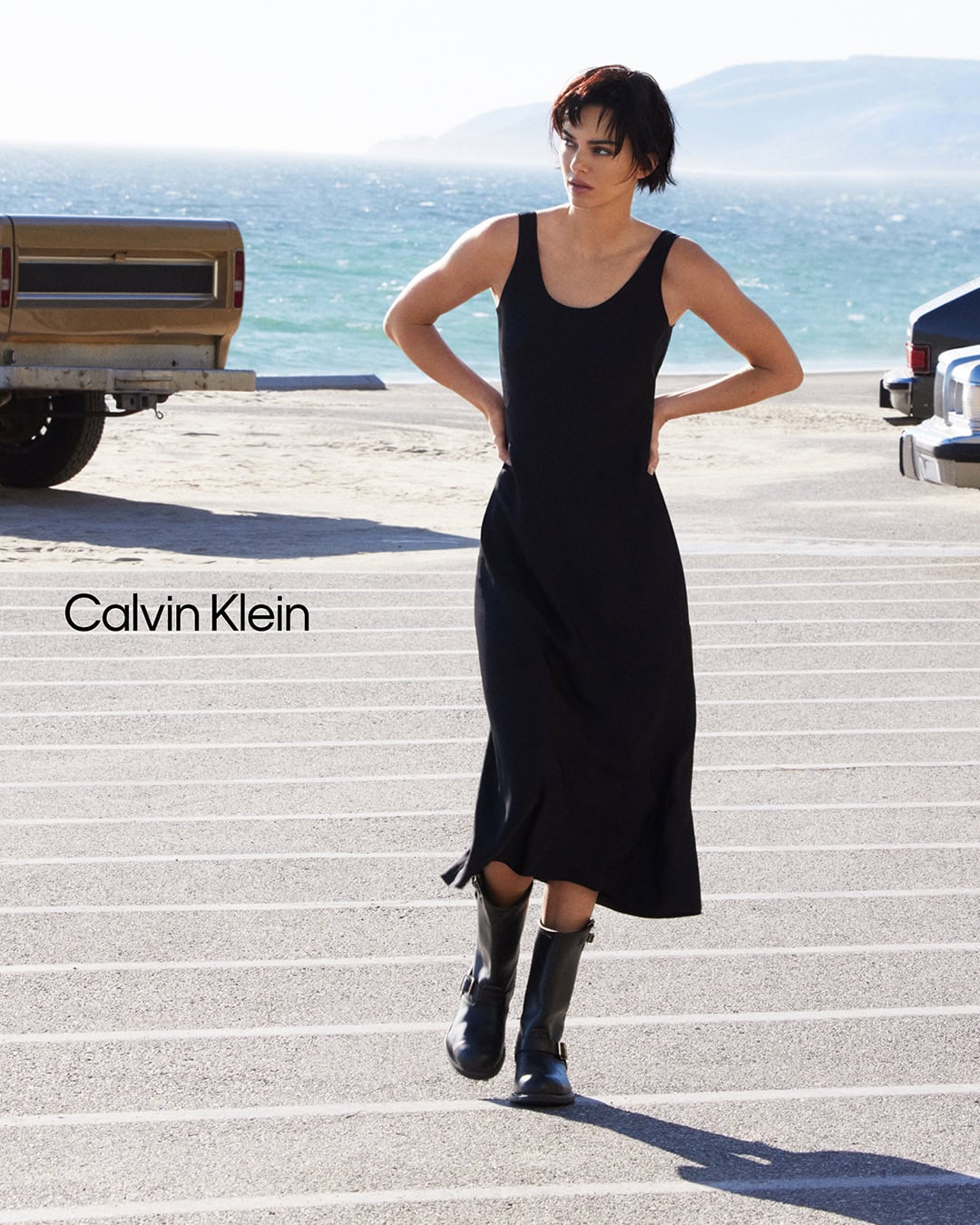 Kendall Jenner in a black slip dress and dark boots walking near a coastal road for Calvin Klein.