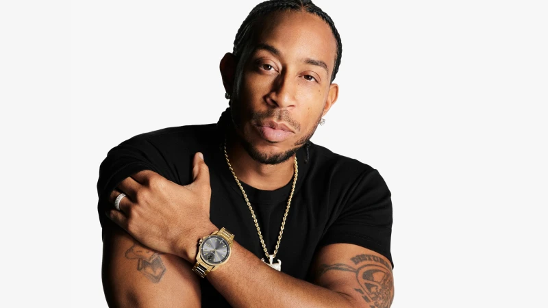 Portrait of Ludacris wearing a gold watch and black t-shirt against a plain white seamless studio background for Movado campaign