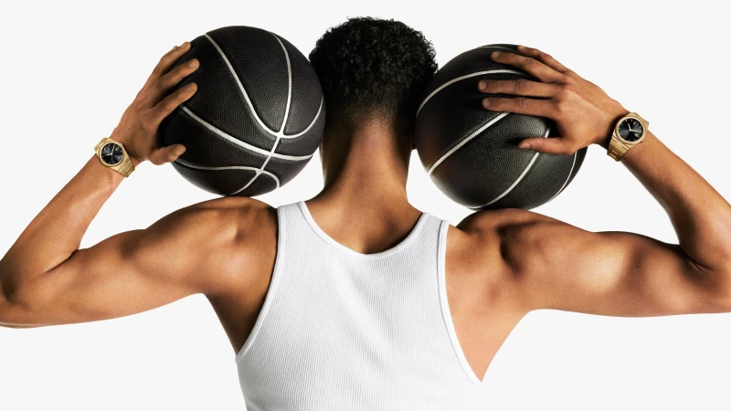 Man facing away holding two black basketballs with gold watches on each wrist against a white background for Movado campaign