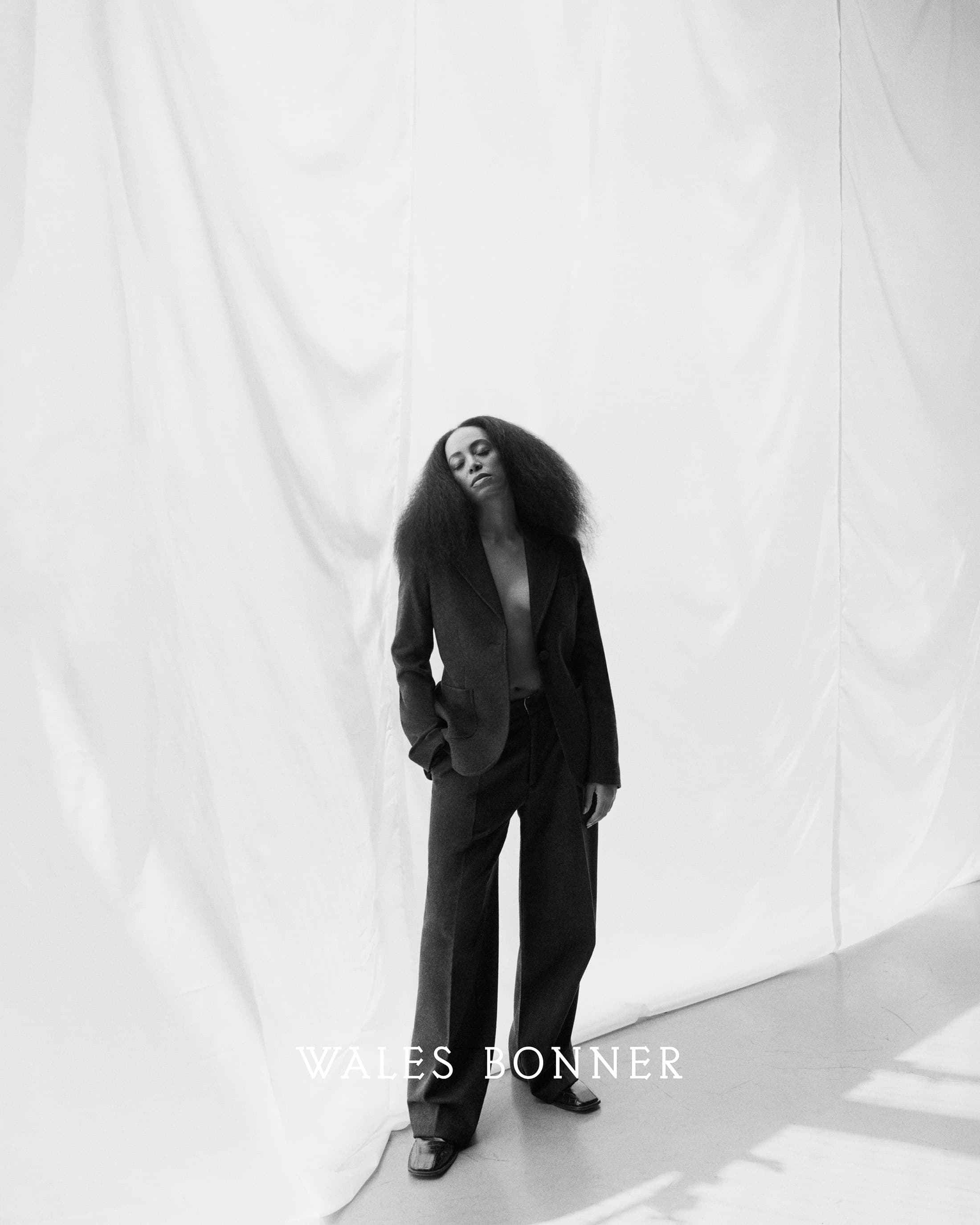 Model with voluminous dark hair wearing a tailored dark blazer and trousers standing against a draped white backdrop.