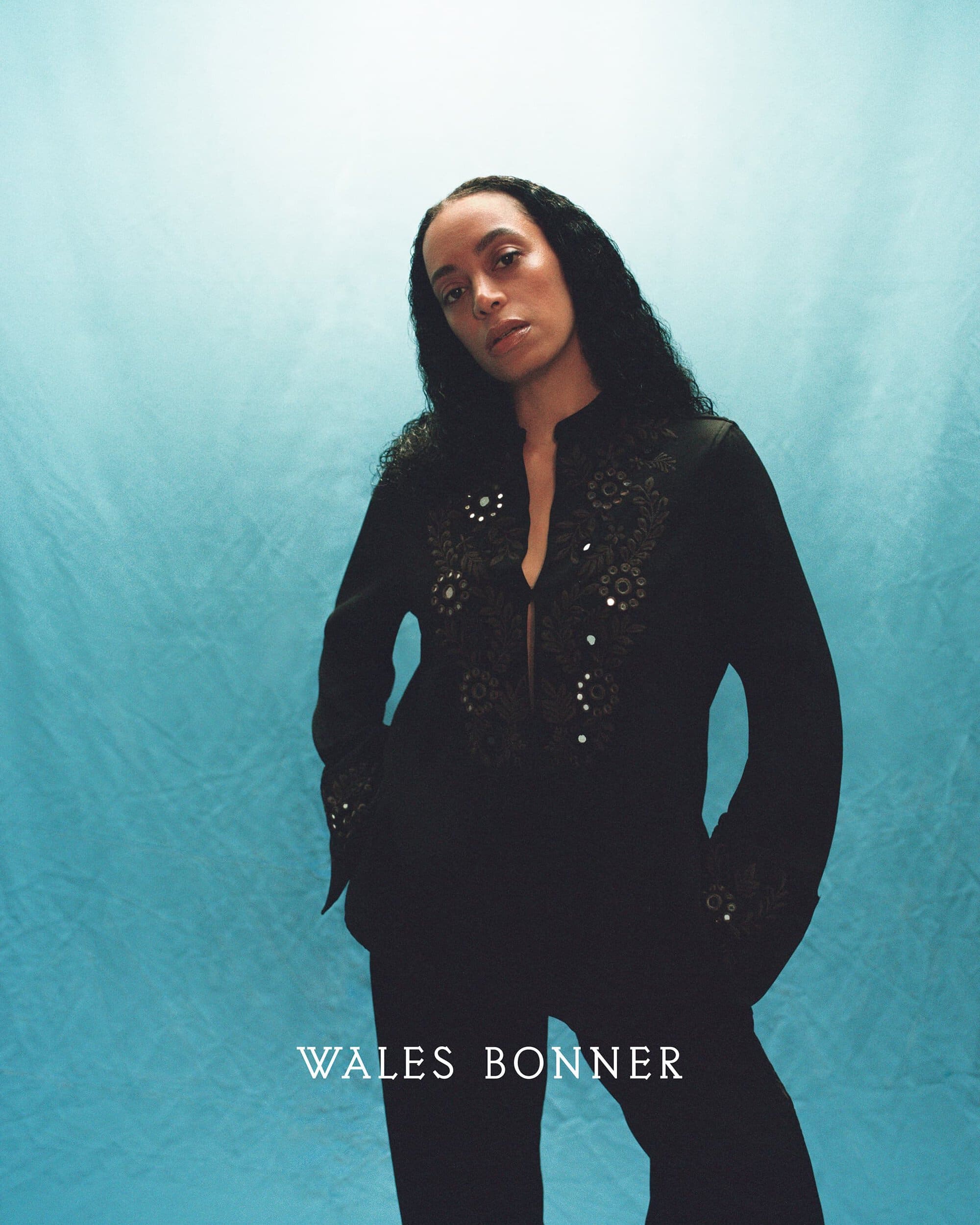 Woman with long dark hair wearing a black embroidered tunic against a blue gradient backdrop for Wales Bonner Fall 2024 campaign