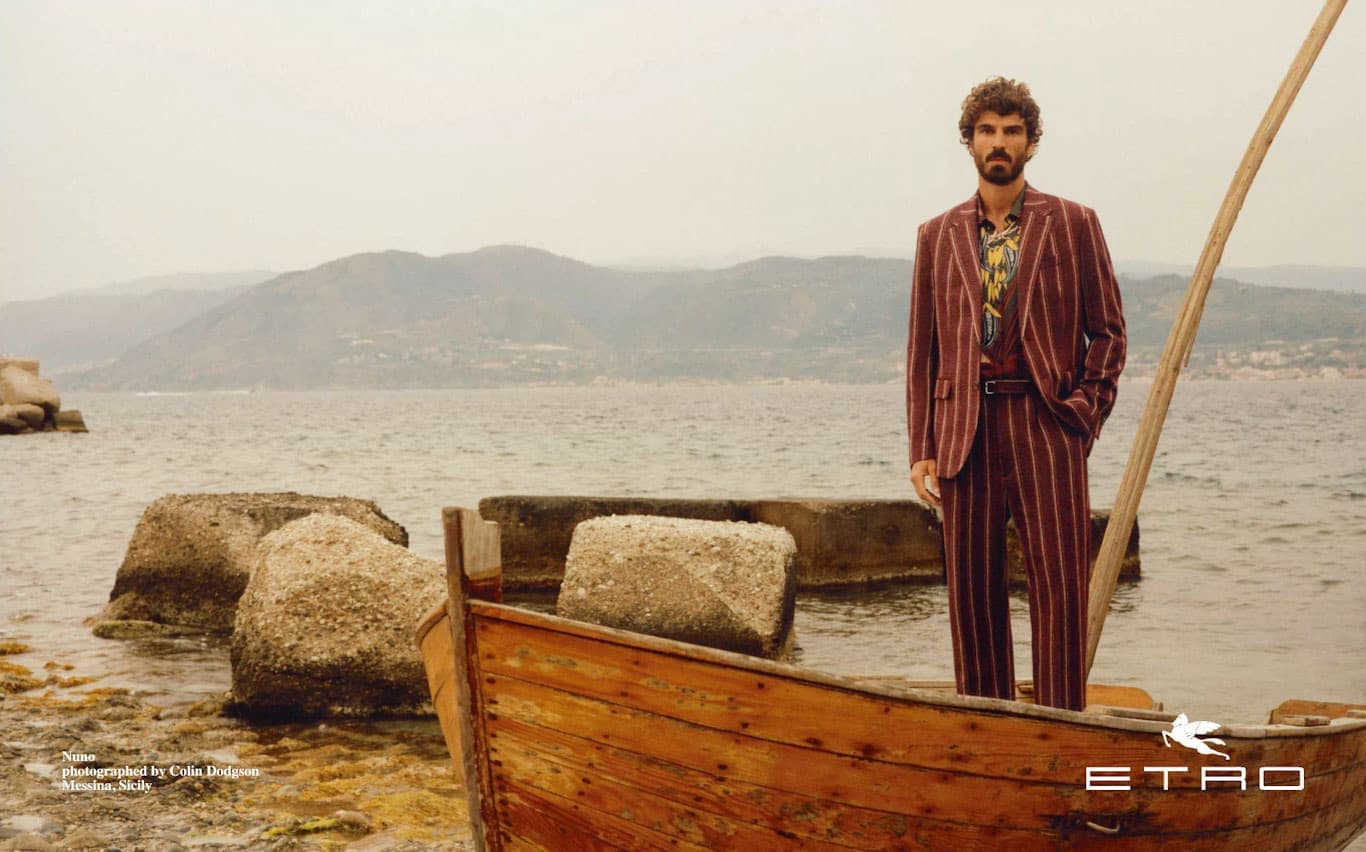 Man standing on a wooden boat by the sea wearing a striped burgundy suit with mountains in the background