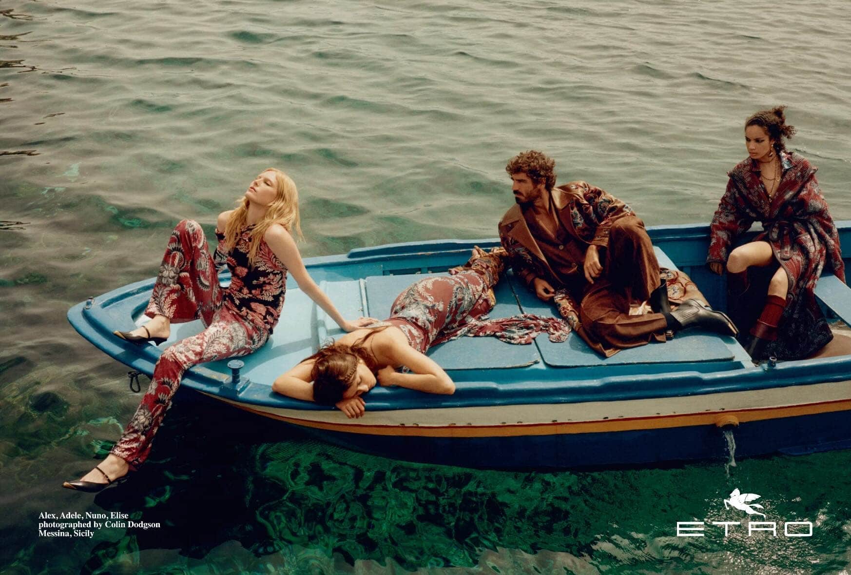 Four models lounging on a blue wooden boat floating on clear green water in Sicily