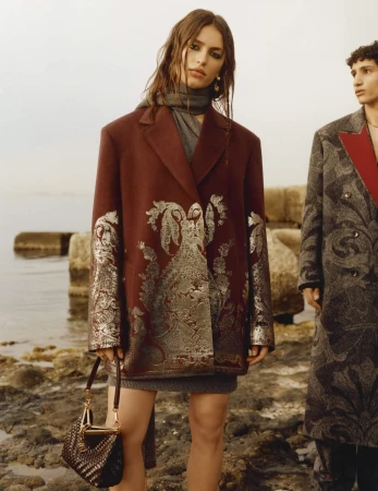 Woman in patterned burgundy blazer holding a handbag standing outdoors near a rocky coastline with a man nearby