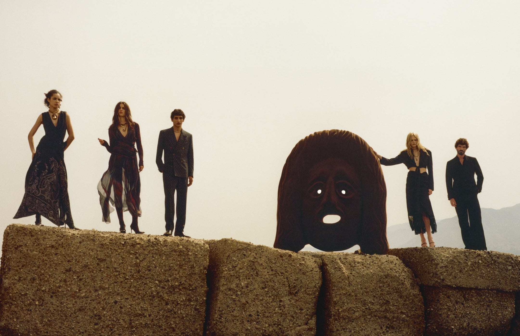 Five models stand atop a stone structure next to a large mask sculpture against an outdoor sky background for Etro Fall 2024 ad campaign.
