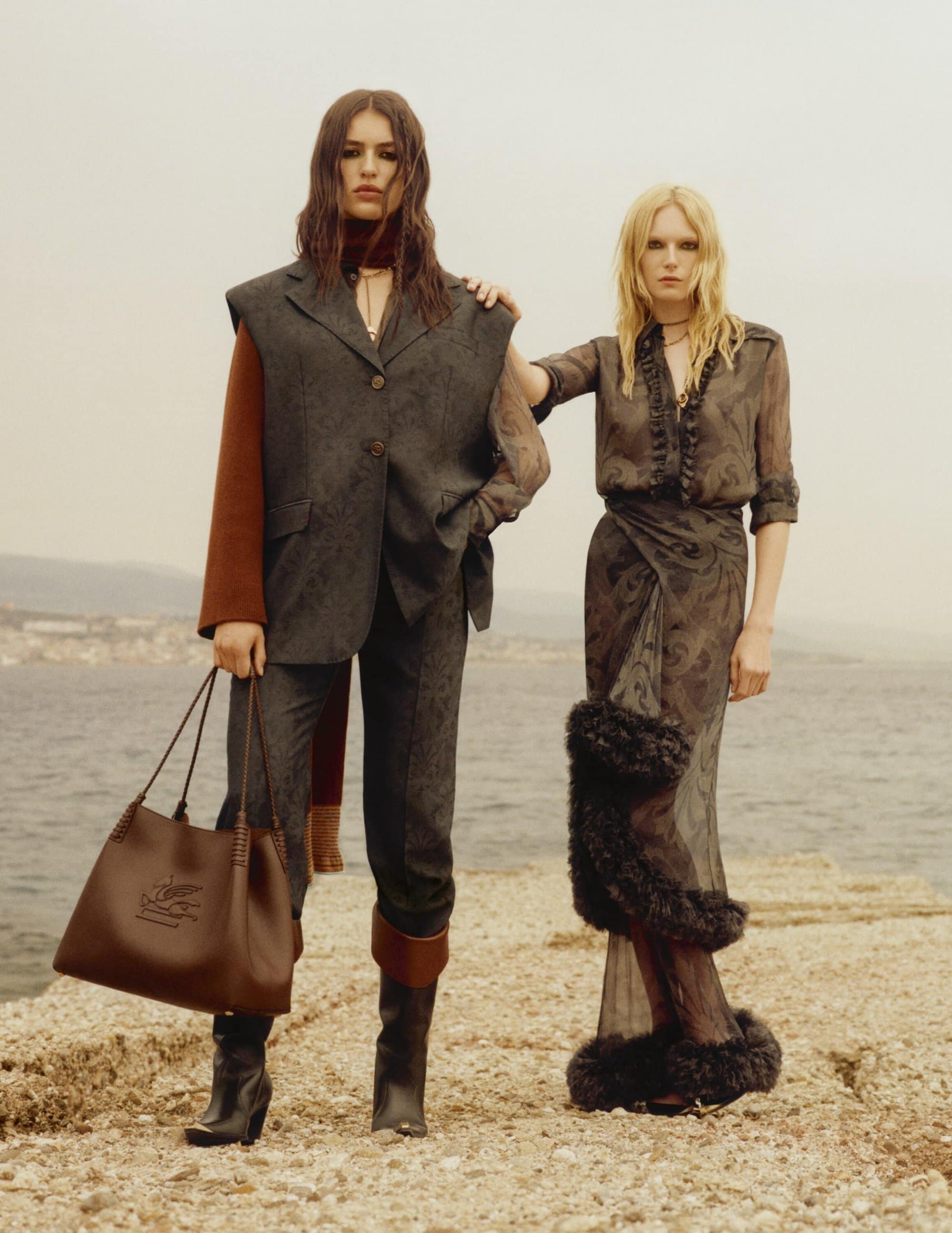 Two models posing outdoors by the sea on a rocky shore wearing patterned dark garments and carrying a leather handbag.