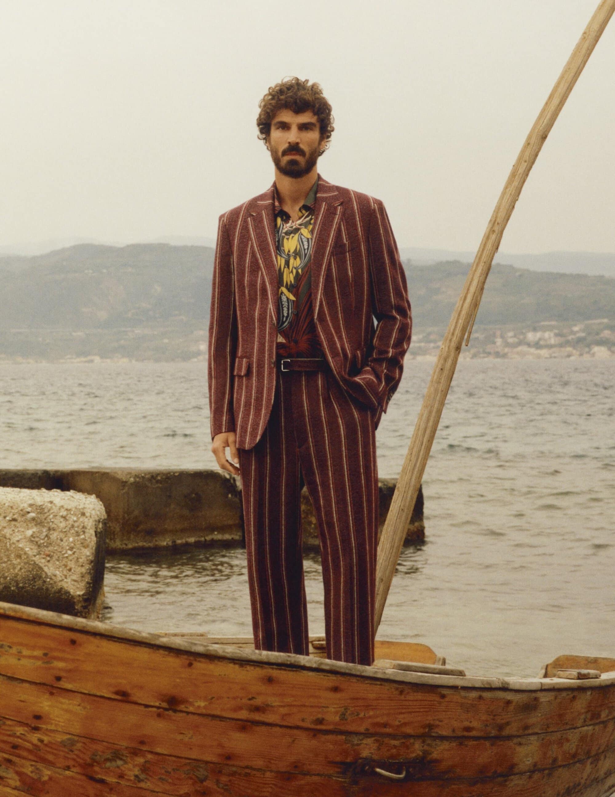 Man wearing a striped burgundy suit standing on a wooden boat by the water with distant hills.