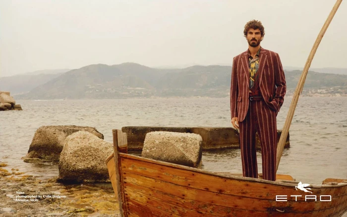 Man standing on a wooden boat by the sea wearing a striped burgundy suit with mountains in the background