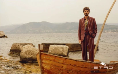 Man standing on a wooden boat by the sea wearing a striped burgundy suit with mountains in the background