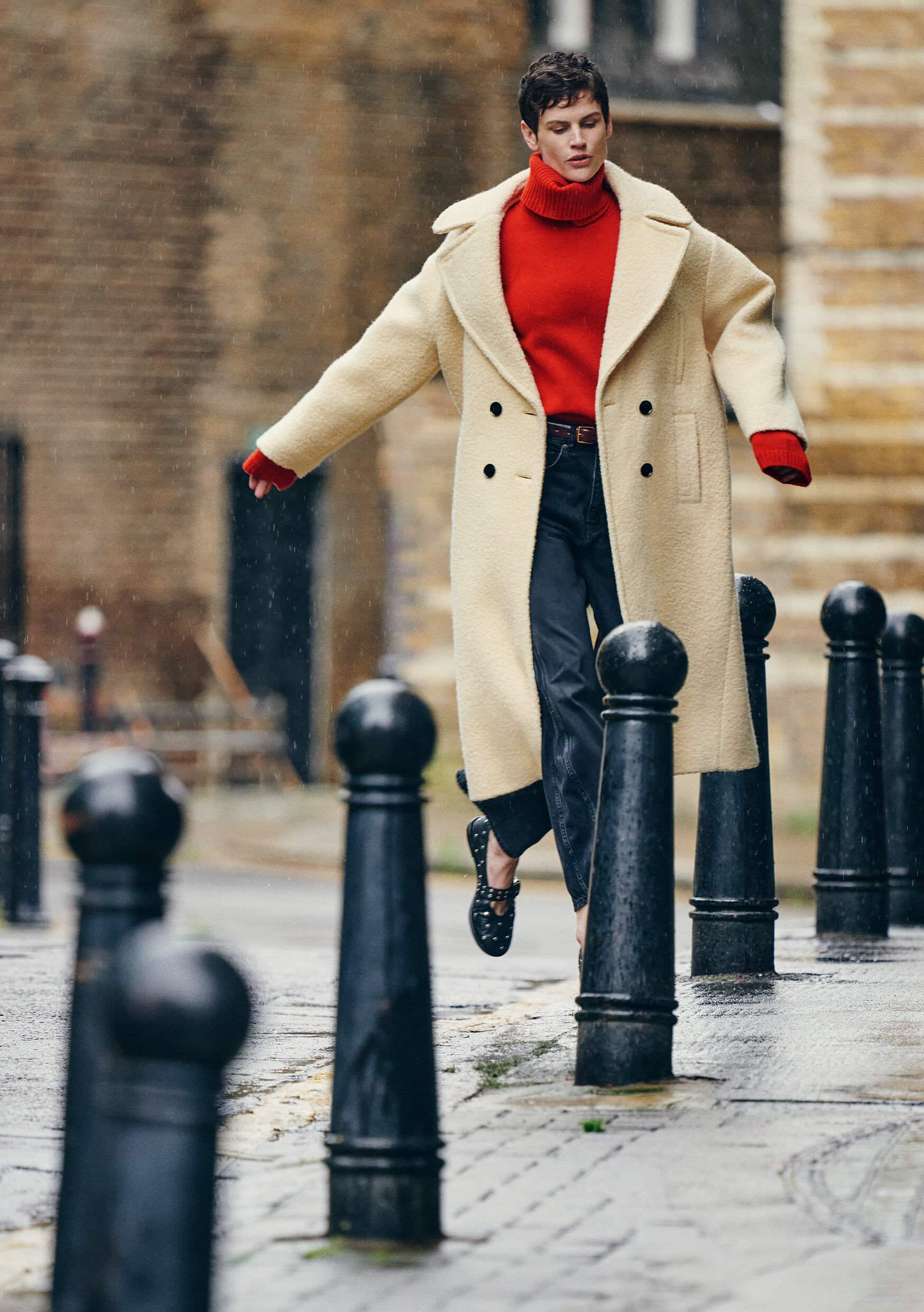 Model Saskia de Brauw wearing a cream coat and red turtleneck walking past bollards on a London street.