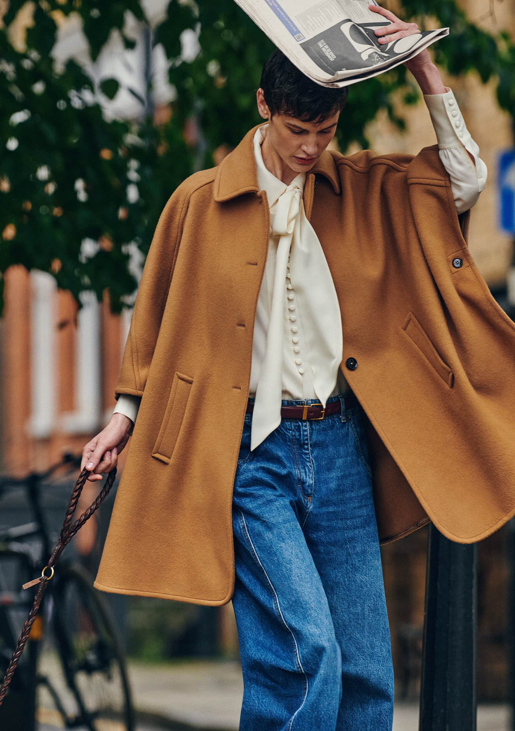 Model in brown wool coat and blue jeans holding newspaper overhead on city street