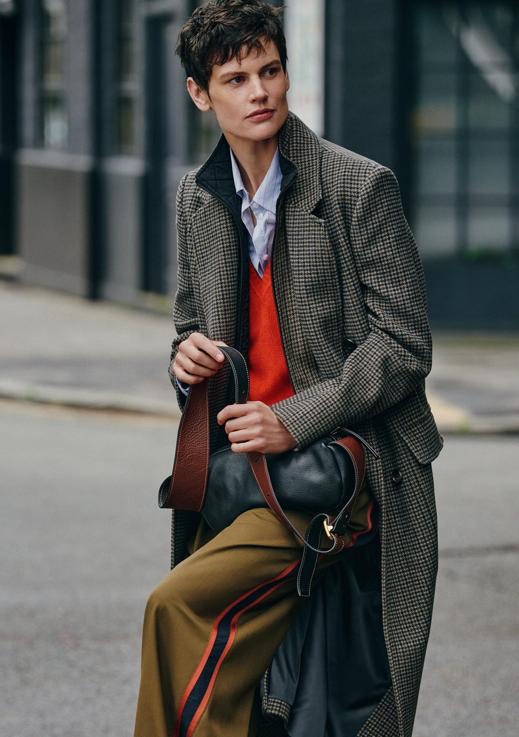 Model Saskia de Brauw wearing a houndstooth coat, red vest and carrying a handbag on an outdoor street
