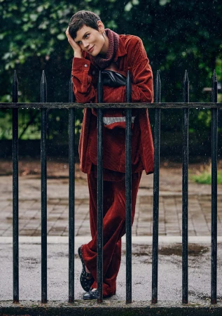 Model Saskia de Brauw in a rust-colored corduroy outfit leaning against black iron railings in a damp urban setting.