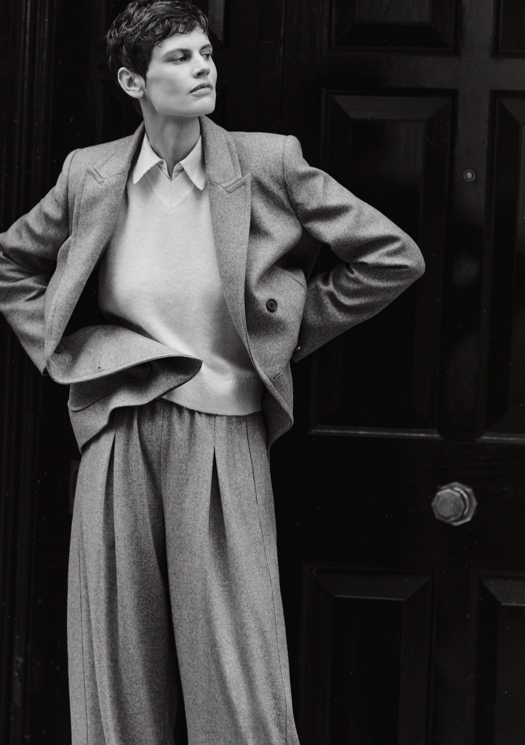 Model Saskia de Brauw in a grey blazer and trousers standing against a dark panelled door