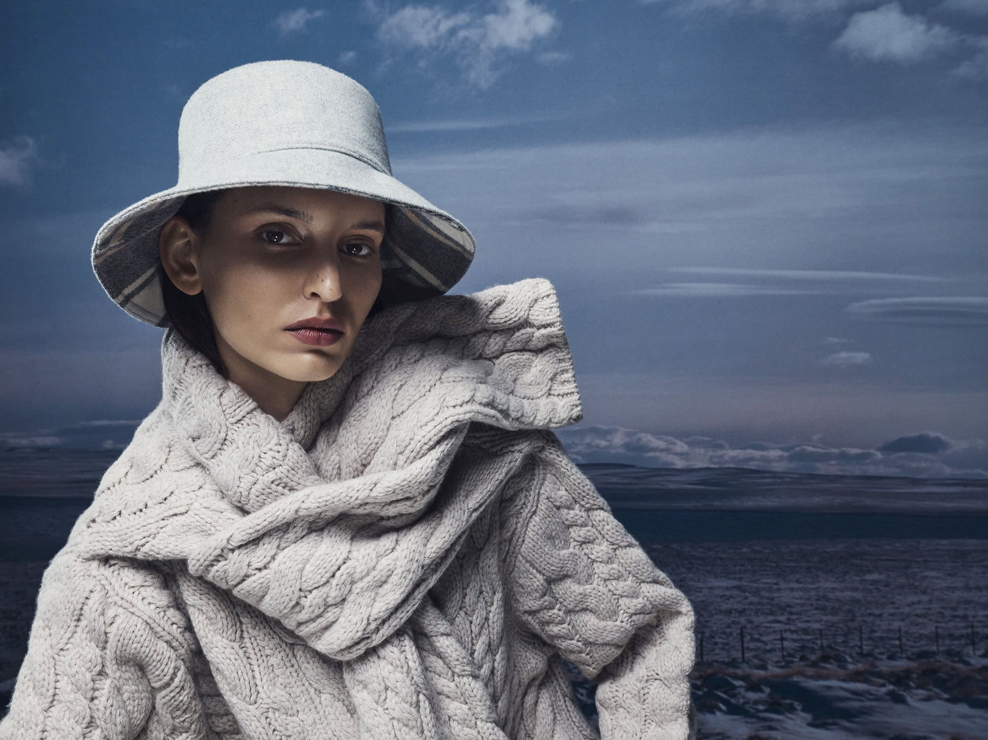 Model wearing a grey bucket hat and textured cable-knit sweater against an overcast sky and landscape background.