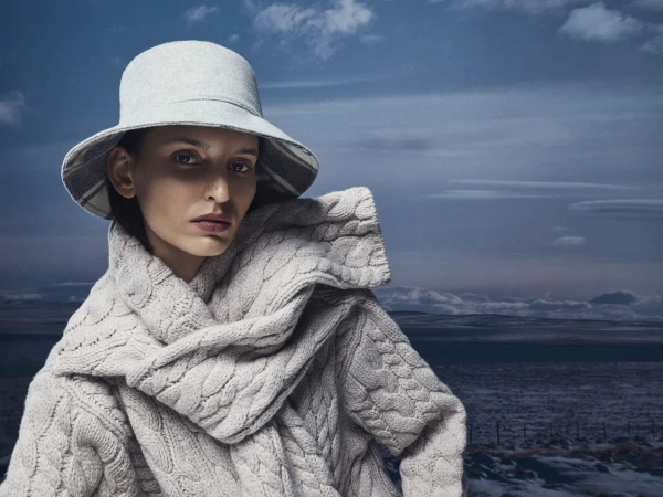 Model wearing a grey bucket hat and textured cable-knit sweater against an overcast sky and landscape background.