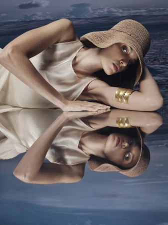 Model wearing a straw hat and gold cuffs reclining on a reflective surface against a moody landscape background.