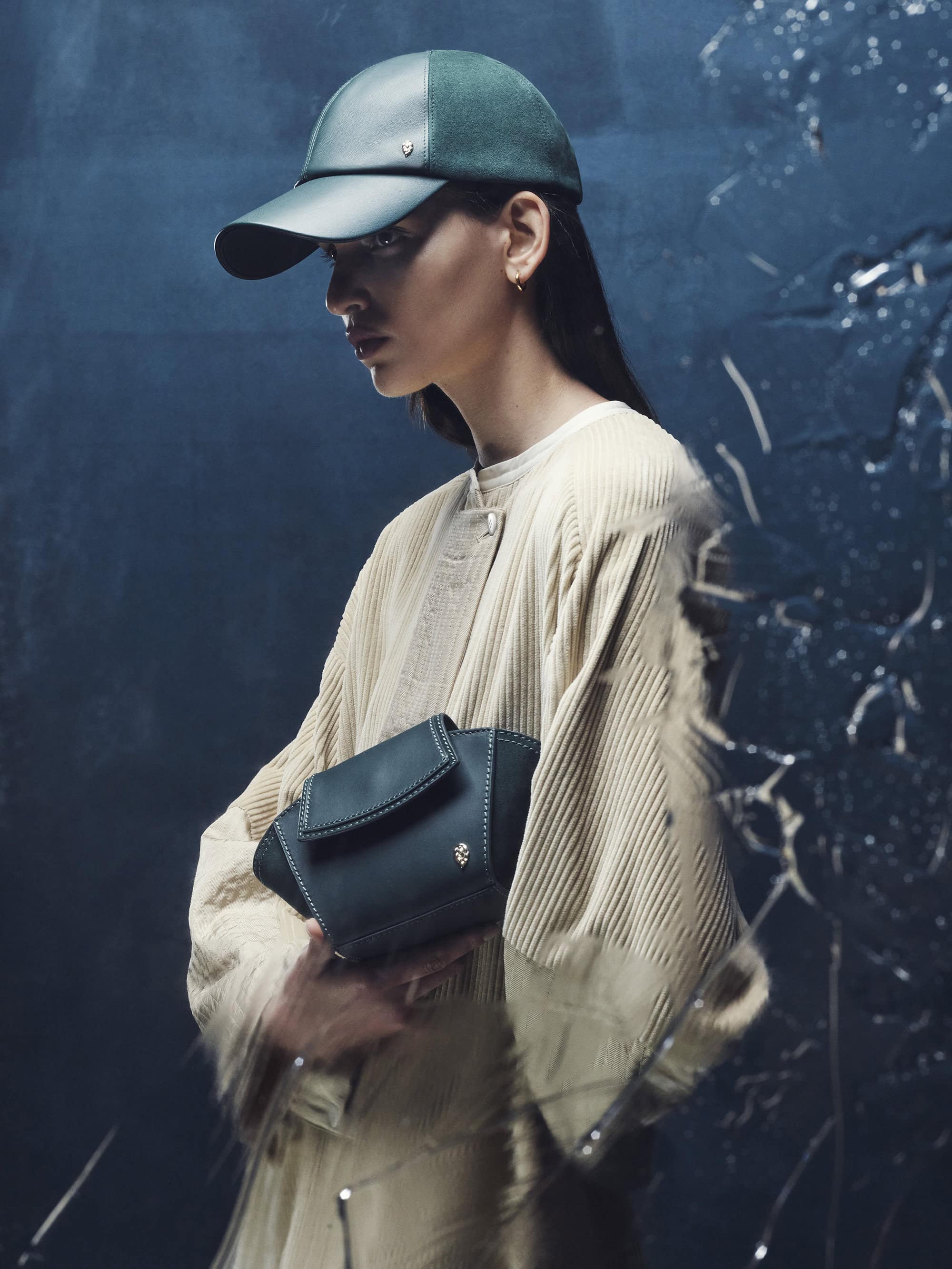 Model wearing a dark green cap and textured cream coat holding a dark bag against a moody blue background