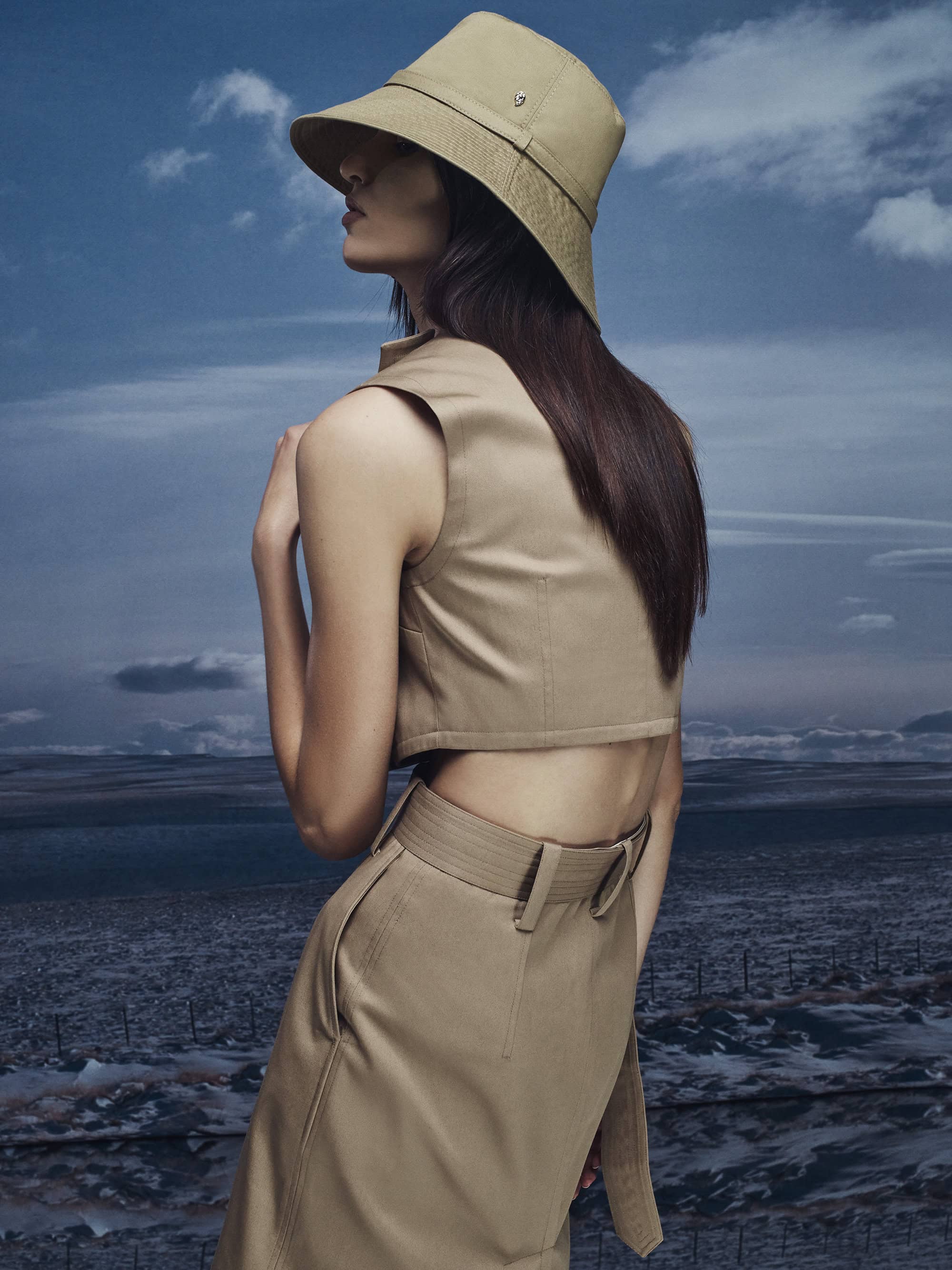 Model wearing a tan bucket hat, sleeveless crop top, and matching skirt against a cloudy sky background for Helen Kaminski