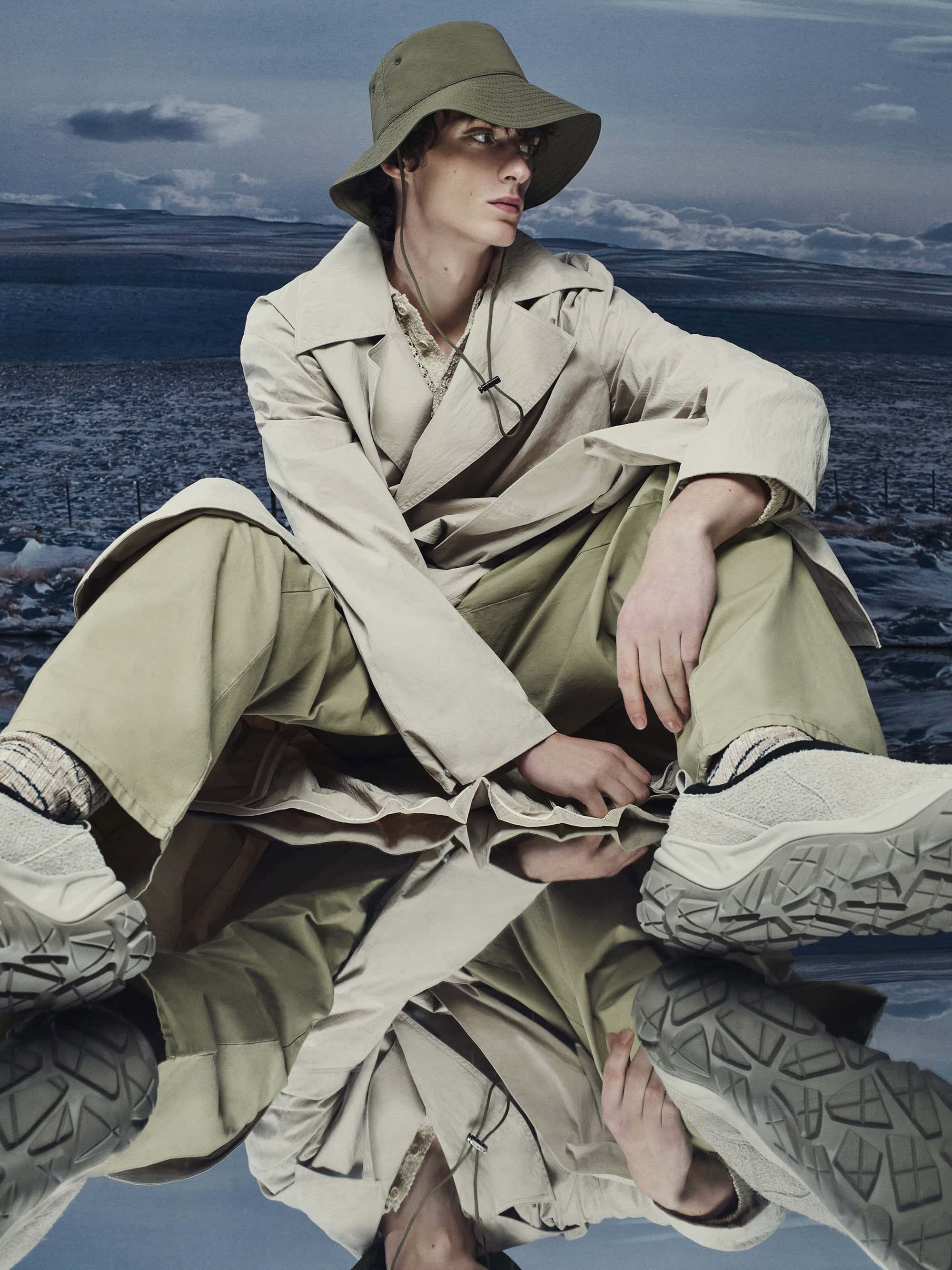 Model wearing an olive bucket hat and neutral trench coat seated on a reflective surface against a cloudy horizon backdrop.
