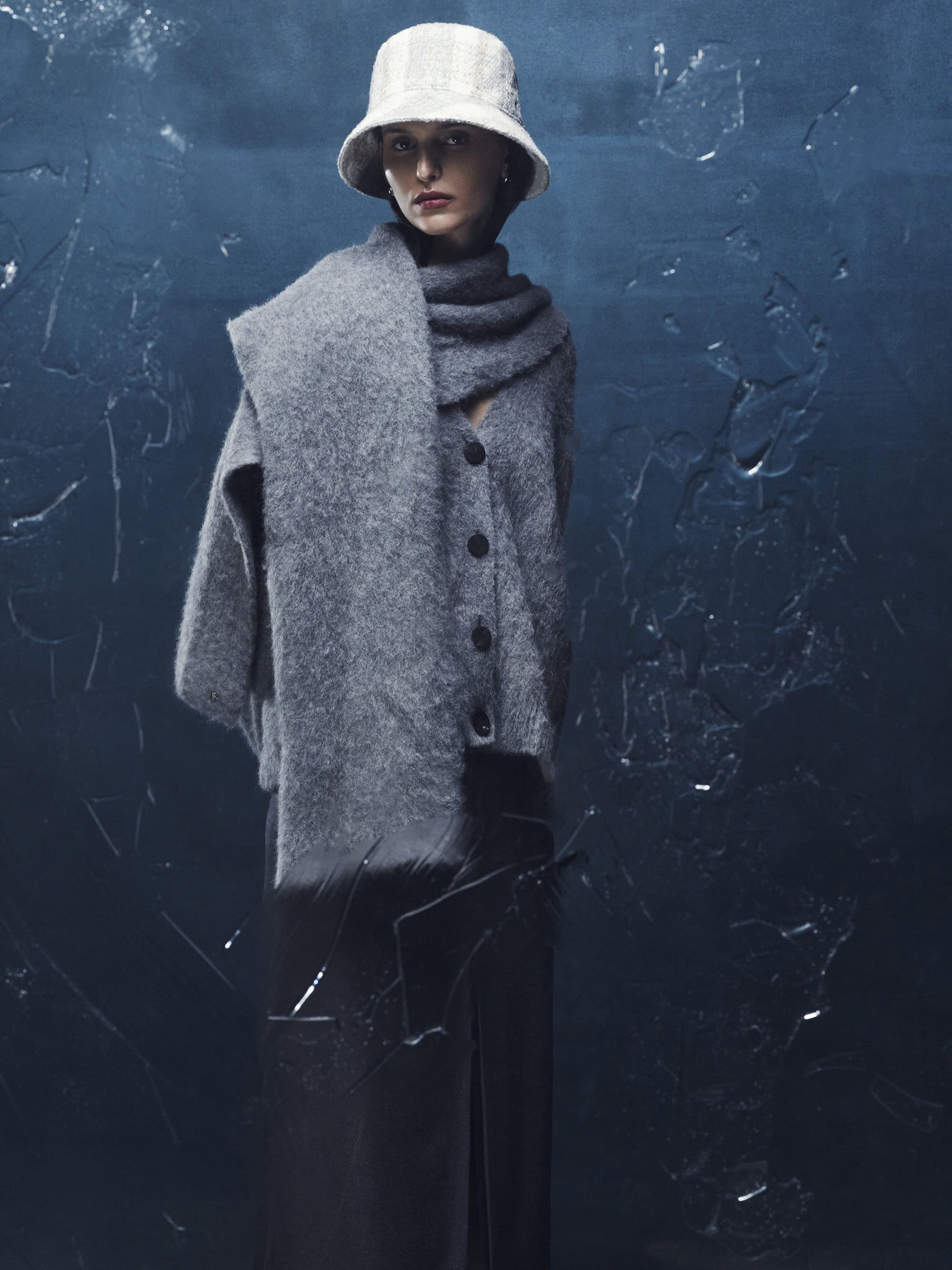 Model wearing a grey wool cardigan, matching scarf, and light bucket hat against a textured dark blue backdrop