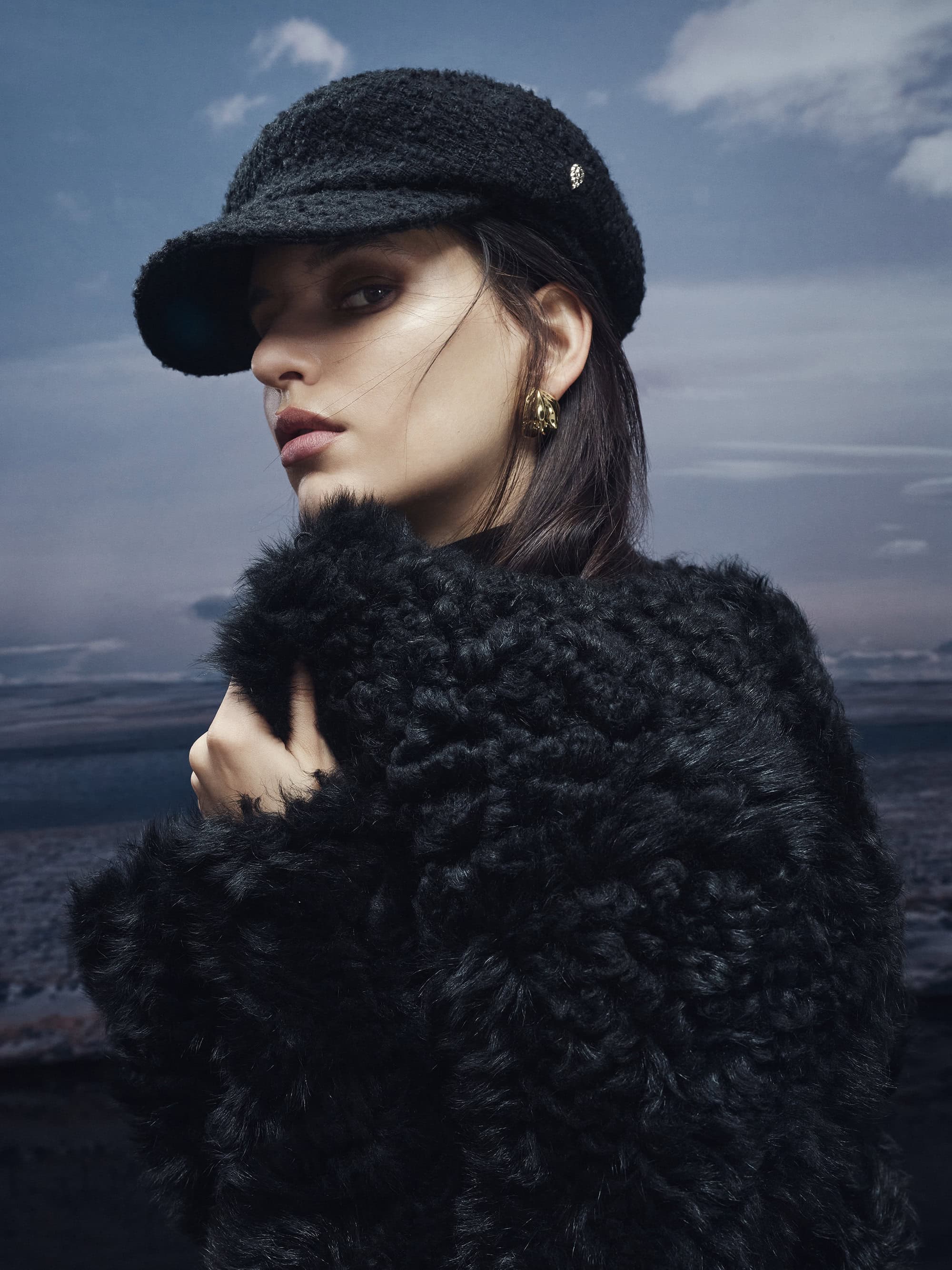 Model wearing a black textured cap and a fluffy black coat against an overcast sky and landscape background