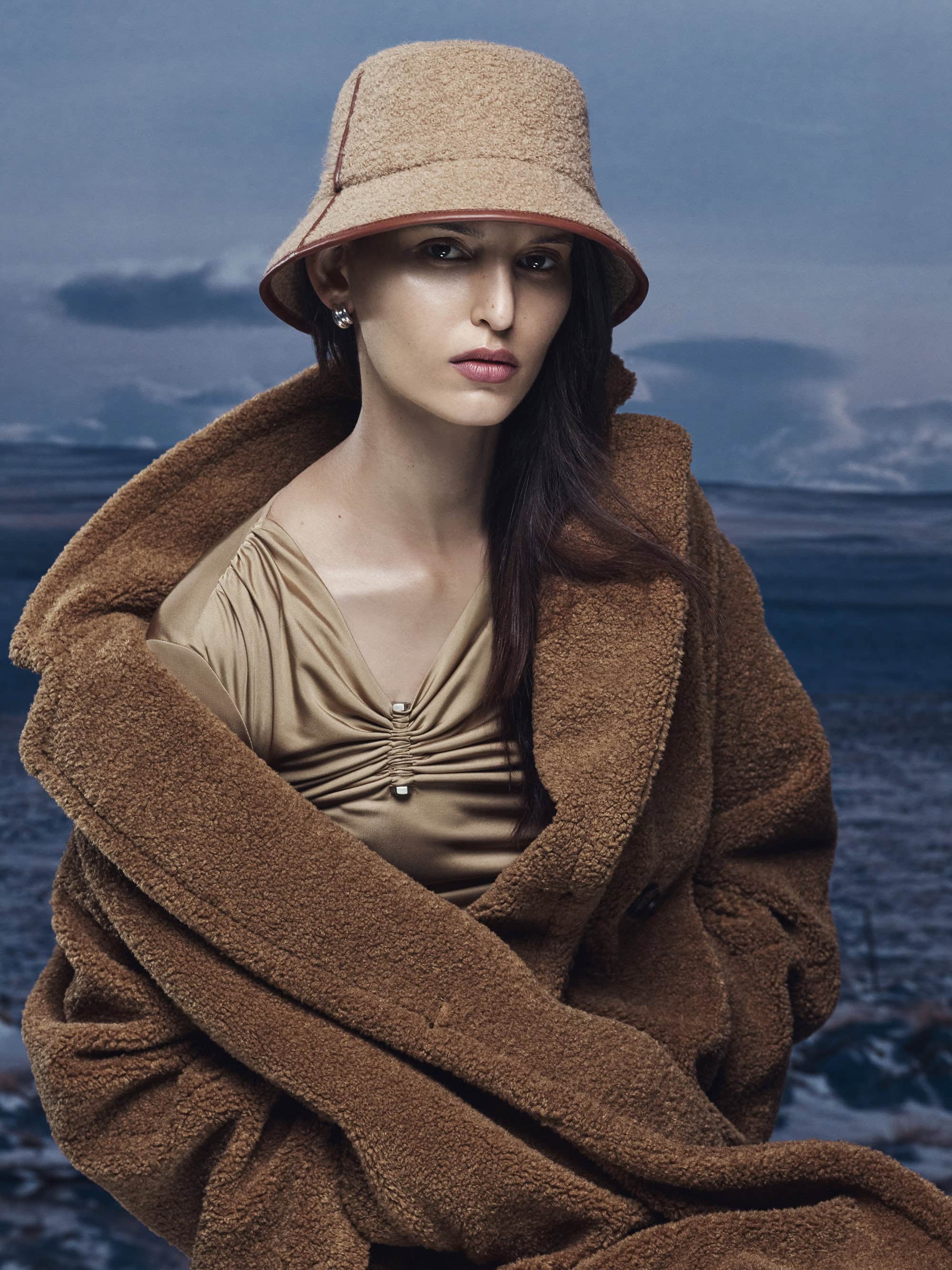 Model wearing a brown bucket hat and textured coat against a dramatic outdoor landscape background