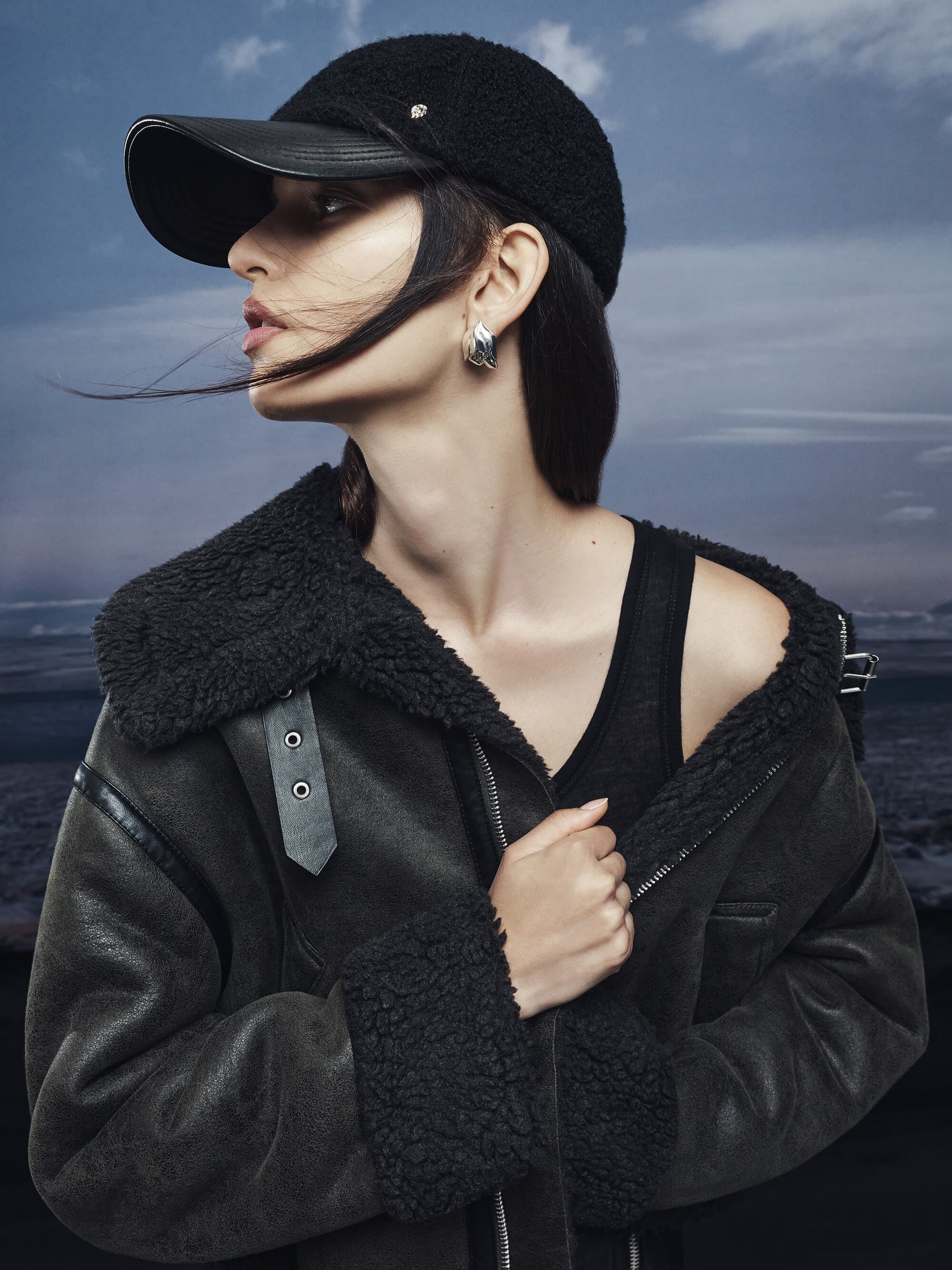 Model wearing a black fleece cap and heavy shearling jacket against an overcast sky and water background