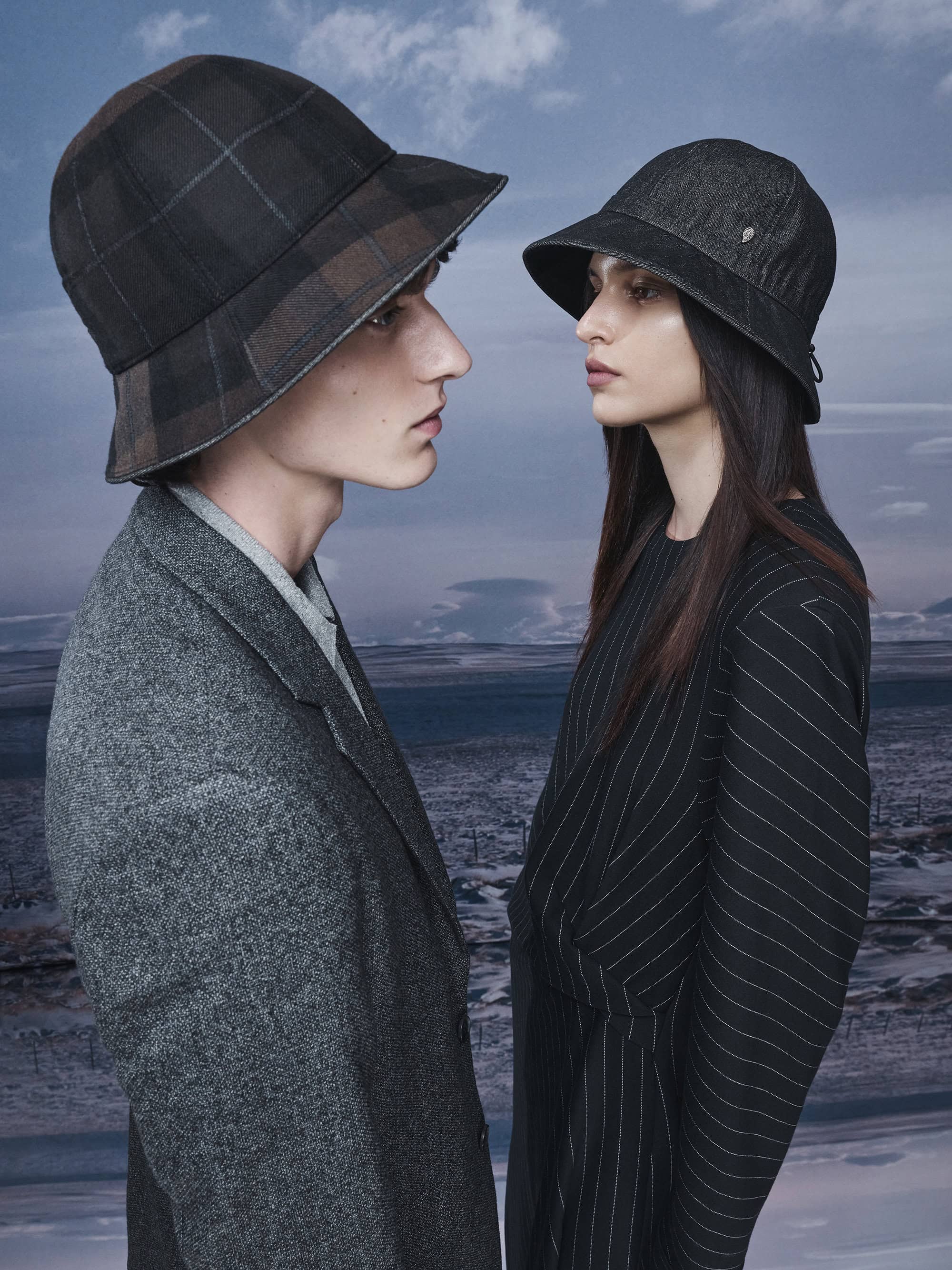 Two models wearing bucket hats and tailored outerwear against an outdoor overcast horizon backdrop