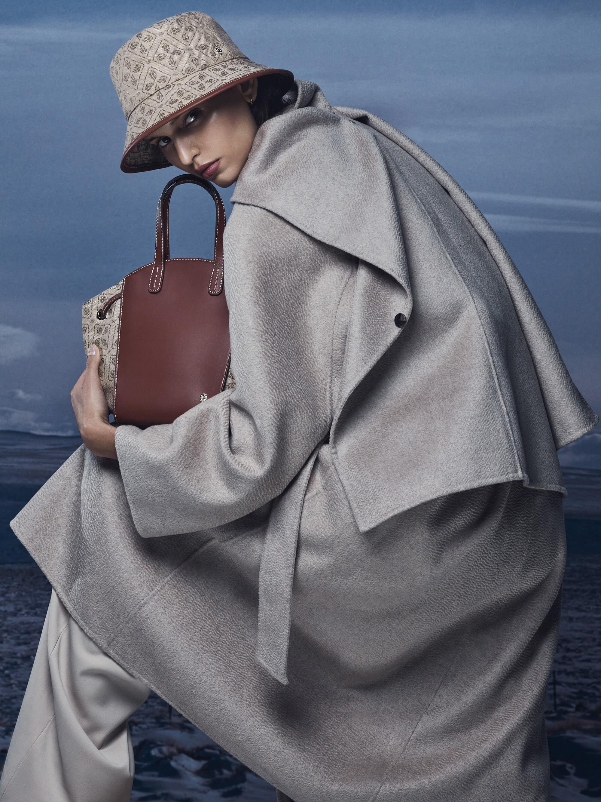 Model wearing a light grey coat and patterned bucket hat holding a brown leather handbag outdoors