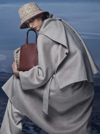 Model wearing a light grey coat and patterned bucket hat holding a brown leather handbag outdoors