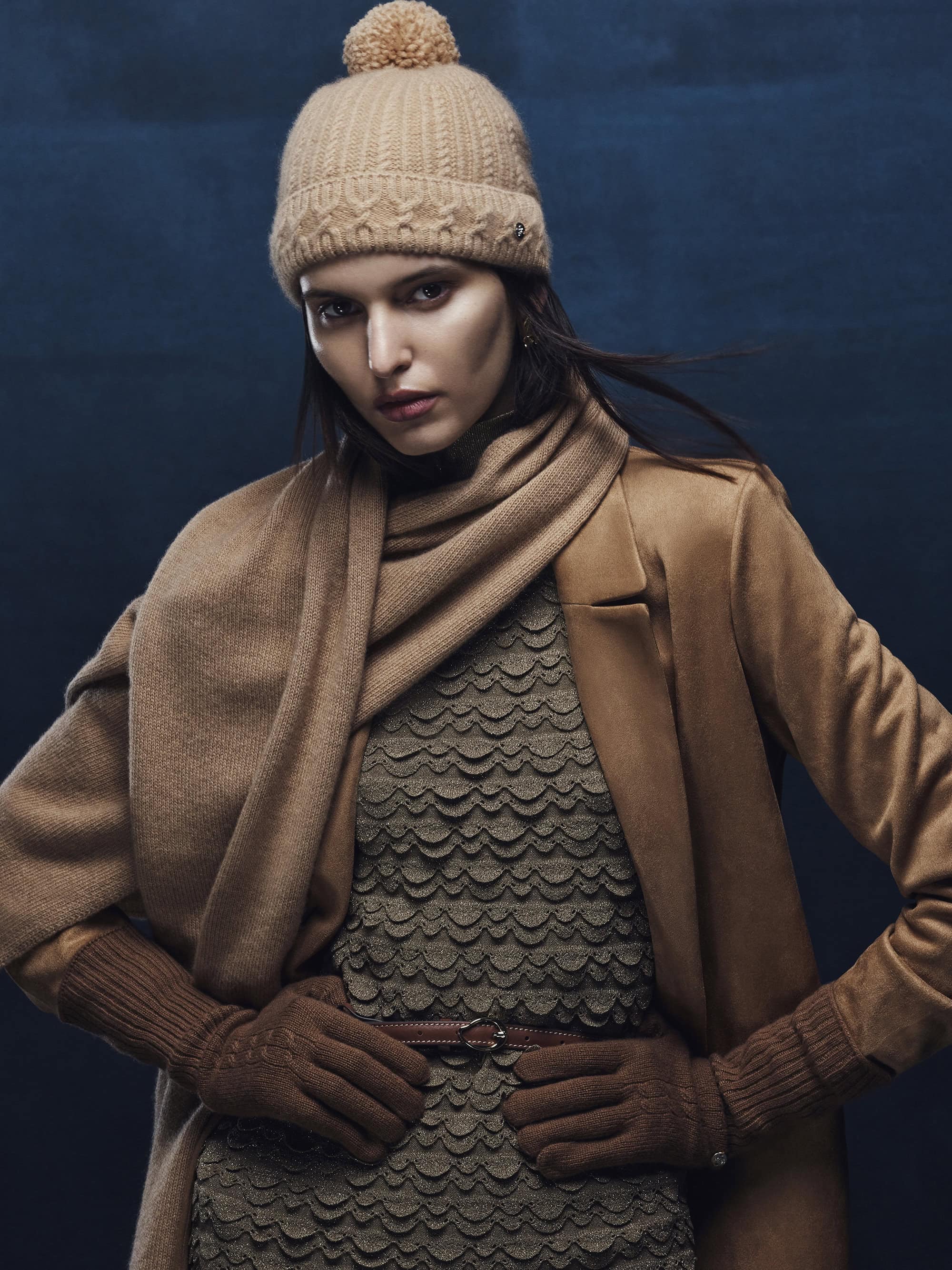 Model in camel knit beanie and matching scarf wearing a tan coat against a dark blue textured background