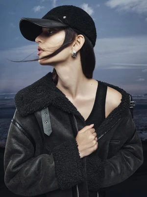 Model wearing a black fleece cap and heavy shearling jacket against an overcast sky and water background