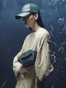 Model wearing a dark green cap and textured cream coat holding a dark bag against a moody blue background