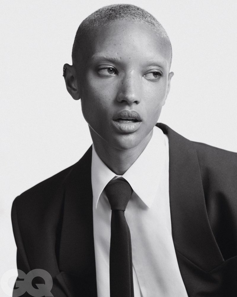 Black and white portrait of a person with short hair wearing a suit and tie, looking off to the side against a plain background.