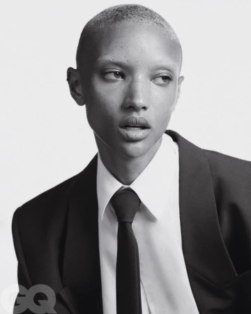Black and white portrait of a person with short hair wearing a suit and tie, looking off to the side against a plain background.