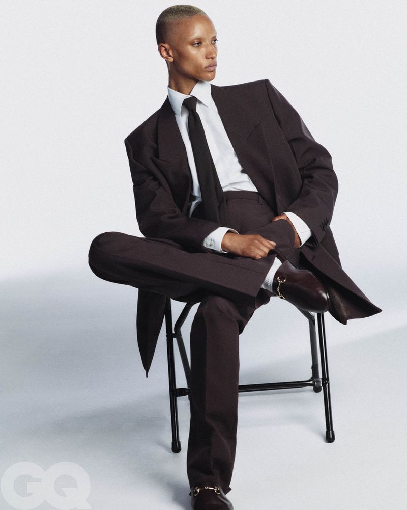 An adult female with a shaved head poses in a brown suit with a white shirt and black tie, seated on a folding chair in a photo studio.