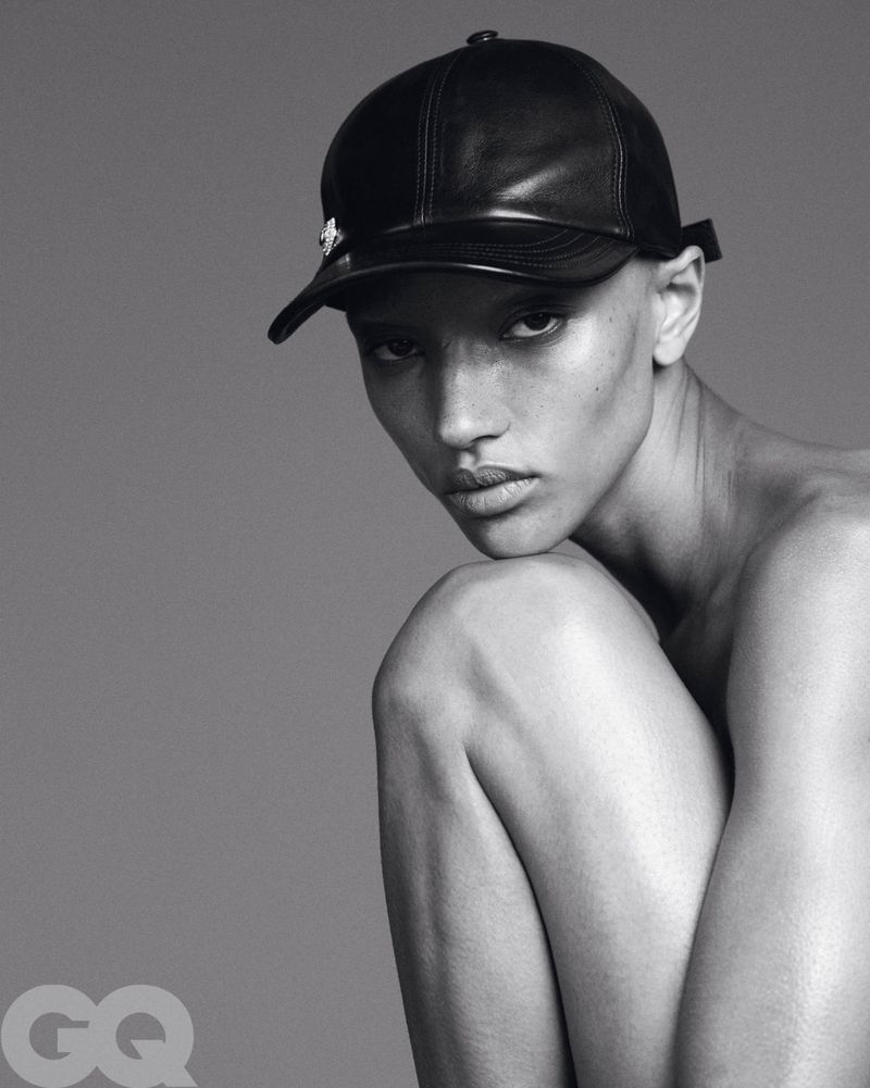 Striking black and white portrait of a model wearing a black leather cap, seated in a studio setting with a minimalist aesthetic.