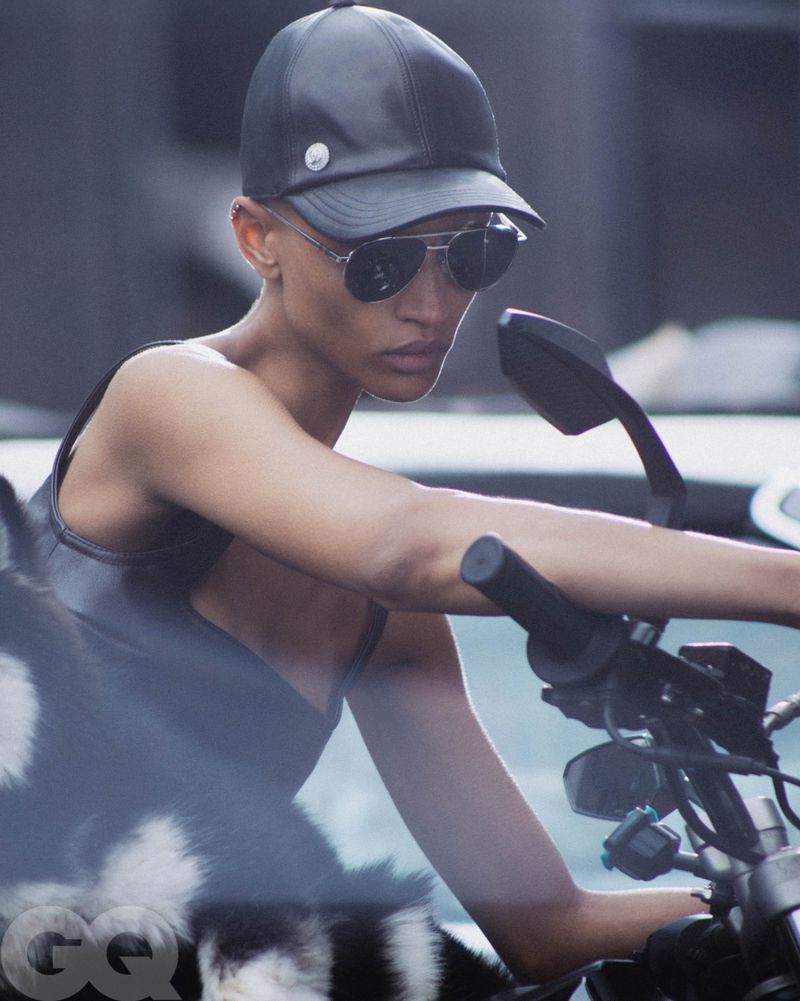 Woman in sunglasses poses on a motorcycle, wearing a black leather cap and top against a blurred urban backdrop, GQ logo in corner.