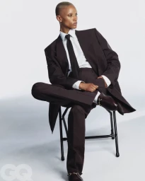 Model Georgia Palmer seated on a chair wearing a dark brown suit, white shirt, and black tie against a white background.