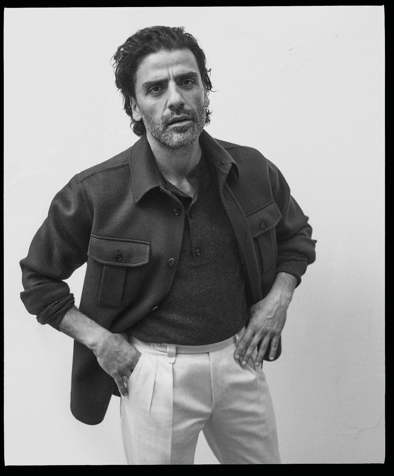 Portrait of Oscar Isaac wearing a dark jacket and light trousers against a neutral white background