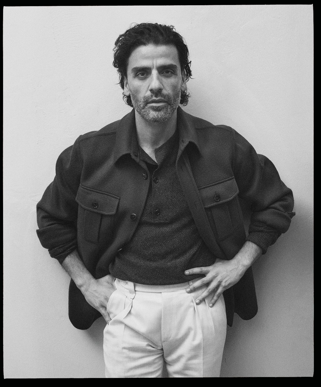 Monochrome portrait of Oscar Isaac wearing a dark collared jacket and light trousers against a plain wall
