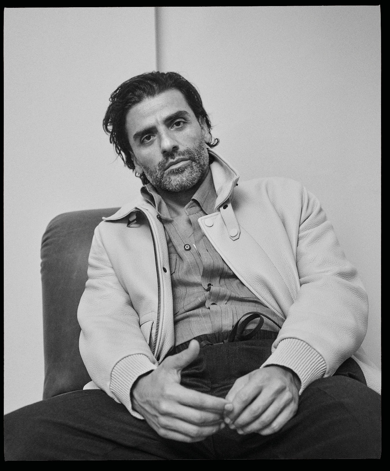 Oscar Isaac wearing a light jacket and collared shirt, seated indoors against a plain background for Brioni.