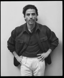 Monochrome portrait of Oscar Isaac wearing a dark collared jacket and light trousers against a plain wall