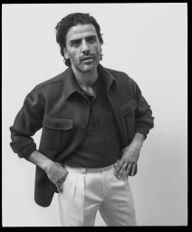 Portrait of Oscar Isaac wearing a dark jacket and light trousers against a neutral white background