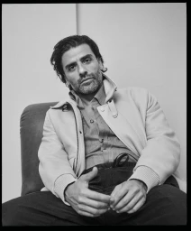 Oscar Isaac wearing a light jacket and collared shirt, seated indoors against a plain background for Brioni.
