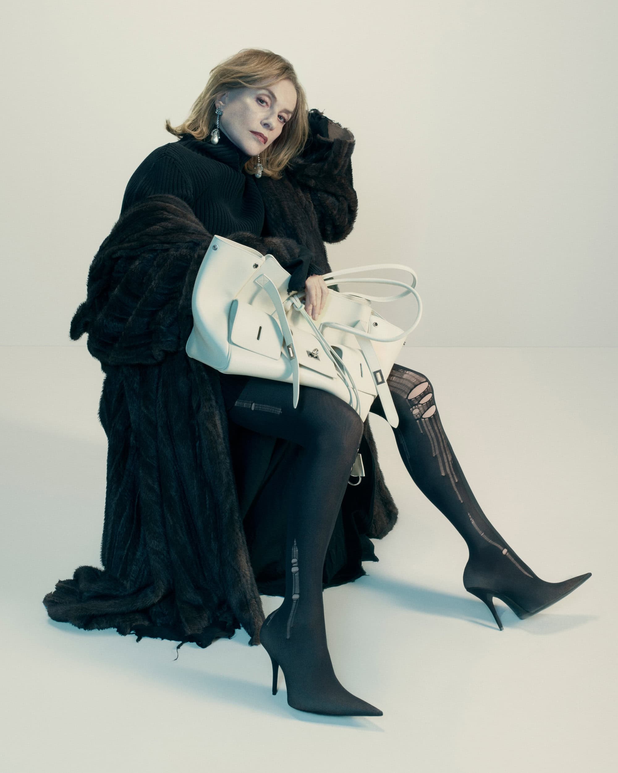 Isabelle Huppert seated in a dark coat holding a large white leather handbag against a plain background for Balenciaga.