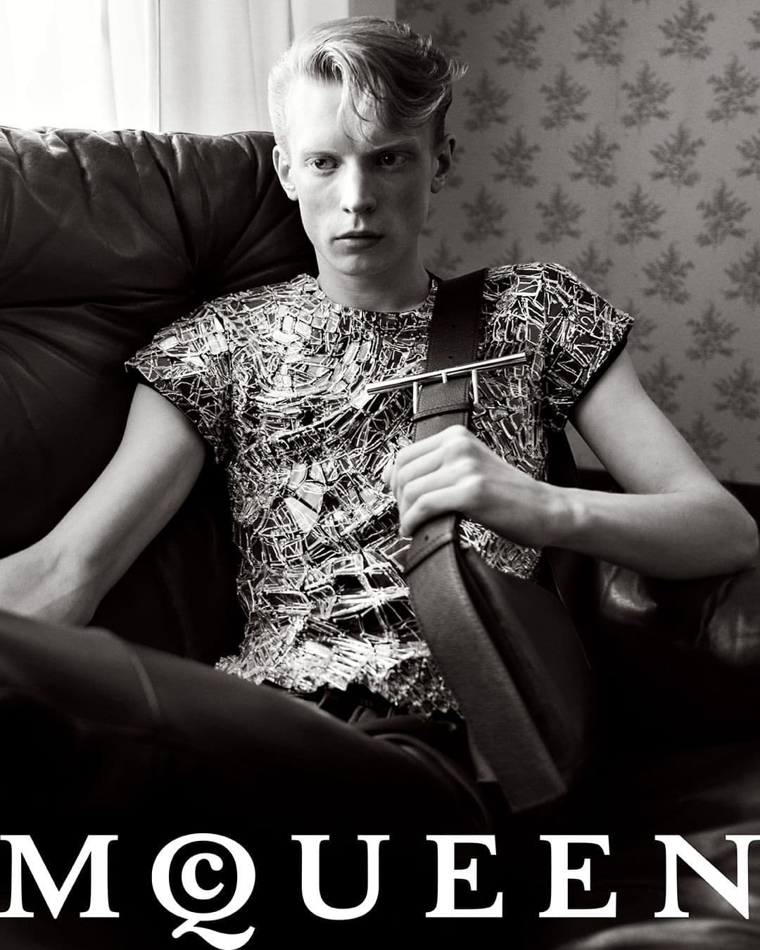 Monochrome portrait of a young man seated in a leather armchair holding a dark leather sling bag for McQueen fall 2024 ad campaign.