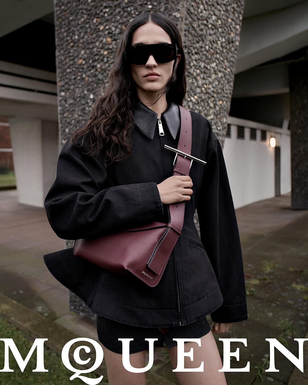 Model wearing dark sunglasses and a black jacket carrying a burgundy crossbody sling bag outdoors.
