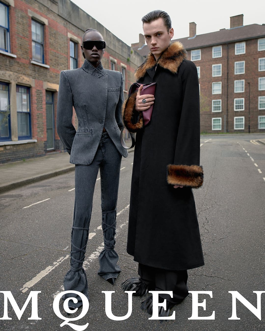 Two models standing on an empty urban street wearing structured grey denim and a long black coat with fur trim for McQueen.